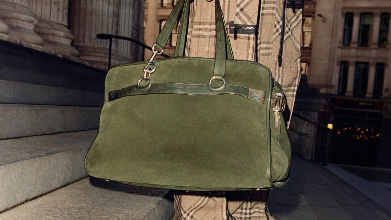 A close-up lifestyle image of a Burberry olive green suede bag photographed outdoors against stone steps and architectural columns, styled with a Burberry check coat. Part of the related content linking to the Burberry Leadership Team section.