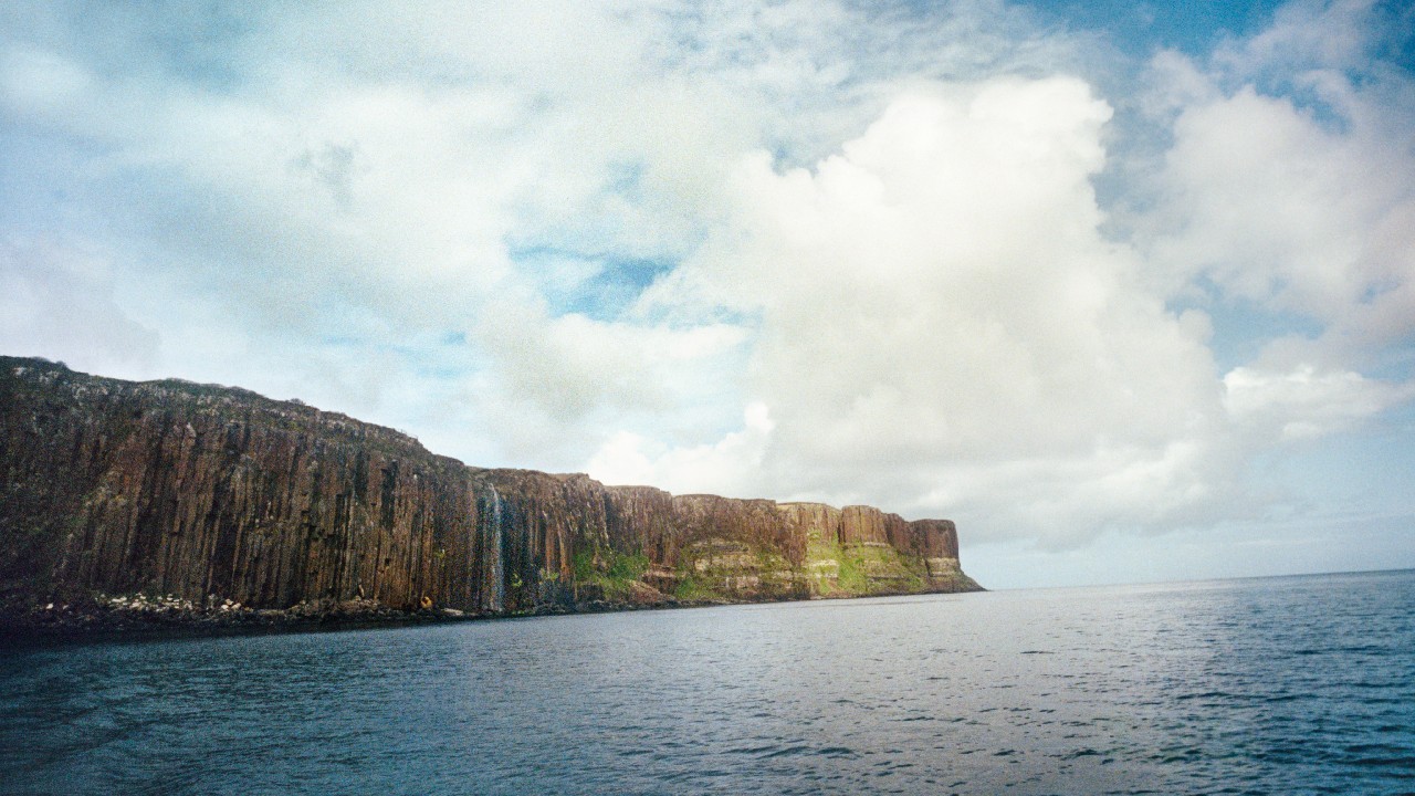 A scenic view of tall coastal cliffs stretching along the shoreline, with calm ocean water and a bright, cloud-filled sky overhead.