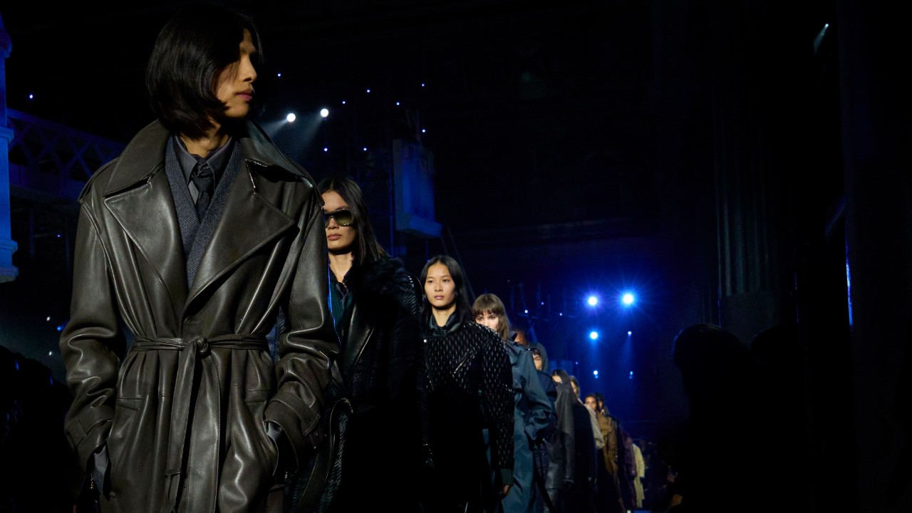 Models wearing dark Burberry outerwear walk down a runway under blue stage lights.