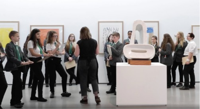 A group of students and a guide gather around a modern sculpture in an art gallery, participating in an educational and interactive cultural experience.