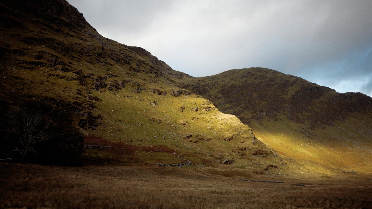 Sunlight breaking through dark clouds over a rugged hillside and grassy valley.