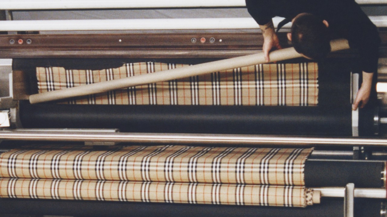 Burberry’s iconic check fabric being processed on industrial rollers inside a textile manufacturing facility.