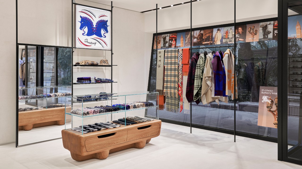 A modern Burberry retail display featuring glass shelving, leather goods, scarves, and apparel arranged in a minimalist boutique setting, representing the brand’s refined craftsmanship and design standards.