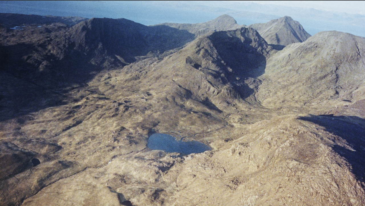A wide aerial view of a rugged mountain landscape with rocky peaks and a small lake nestled among the slopes.
