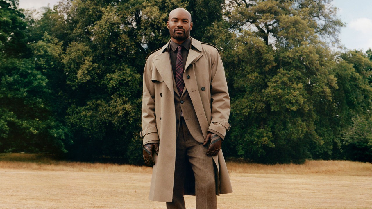Man standing outdoors in a classic Burberry trench coat in a field with trees behind him.