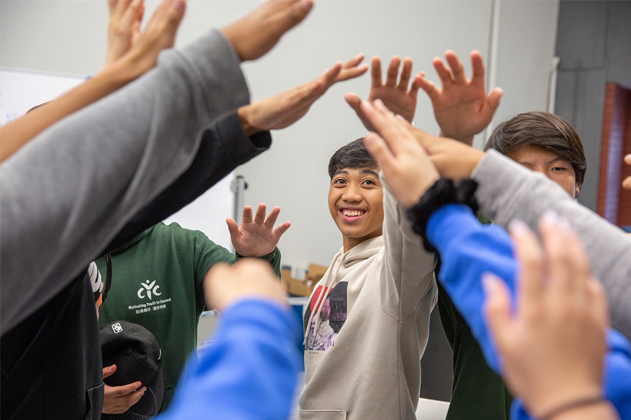 A group of young people standing together in a classroom environment, raising their hands in a collective high-five gesture, symbolizing youth empowerment and collaboration.