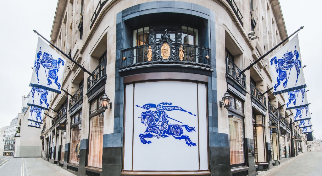 An exterior view of the Burberry flagship store adorned with blue Equestrian Knight Design flags, showcasing the brand’s heritage and visual identity along a London streetscape.