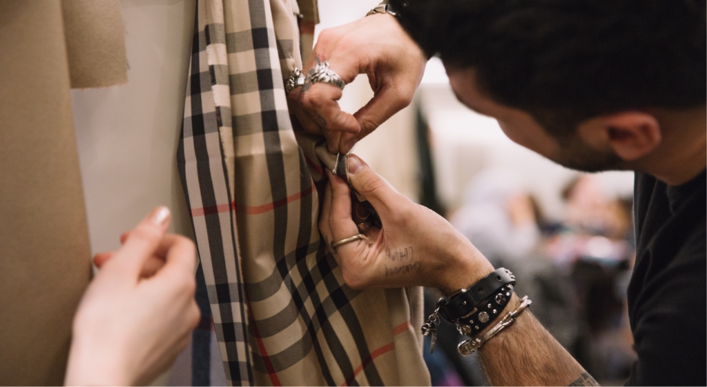 Close-up image of a craftsperson adjusting Burberry check fabric during a tailoring or garment-making process. The hands, tools, and iconic pattern highlight the brand’s focus on craftsmanship and detail.