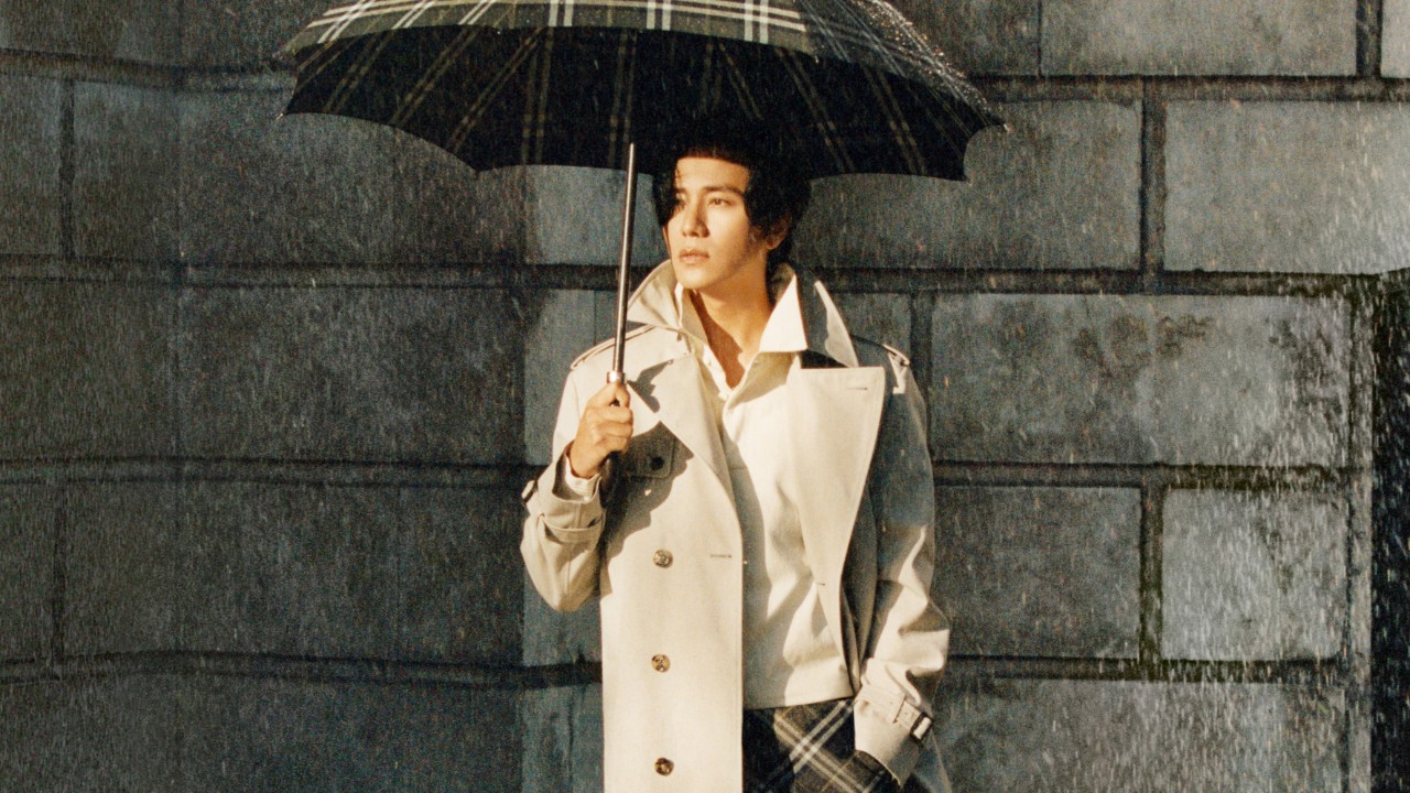 A person wearing a Burberry trench coat walks through a rainy urban setting while holding an umbrella. 