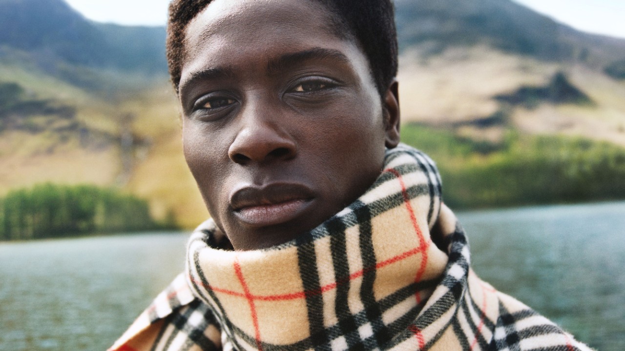 A person wearing a checked Burberry scarf stands outdoors near a lake, with hills and greenery in the background, representing Burberry’s commitment to diversity, equity and inclusion.